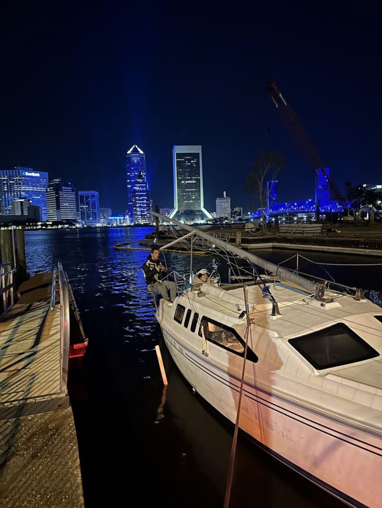 Docked boat in a brightly illuminated Jacksonville waterfront at night, with two people aboard, showcasing the city's skyline and vibrant colors.