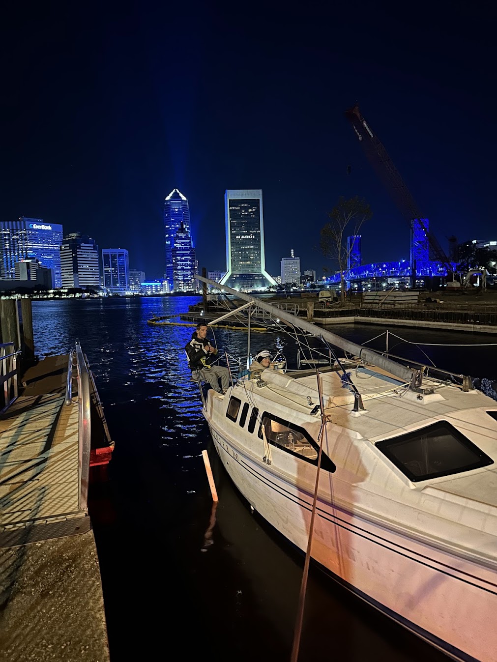 Docked boat in a brightly illuminated Jacksonville waterfront at night, with two people aboard, showcasing the city's skyline and vibrant colors.