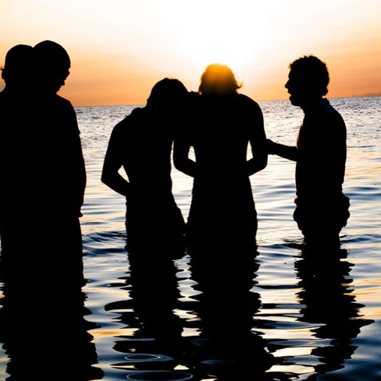 Silhouetted figures stand in shallow water at sunset, engaged in conversation. The tranquil scene captures a moment of connection against a colorful horizon.