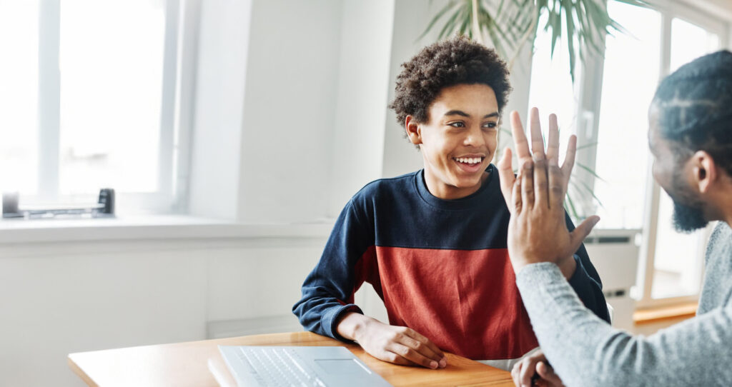 A teenage boy smiles and shares a high-five with an adult, promoting connection and positive interaction in a bright, modern workspace.
