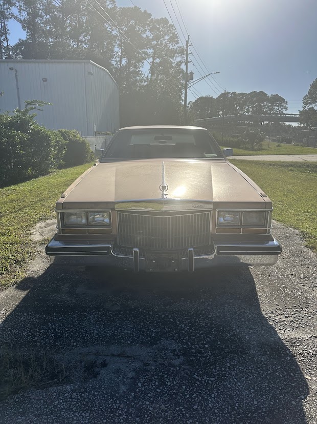 Front view of a vintage Cadillac, showcasing its distinctive chrome grille and beige exterior, parked on a gravel surface with greenery nearby.