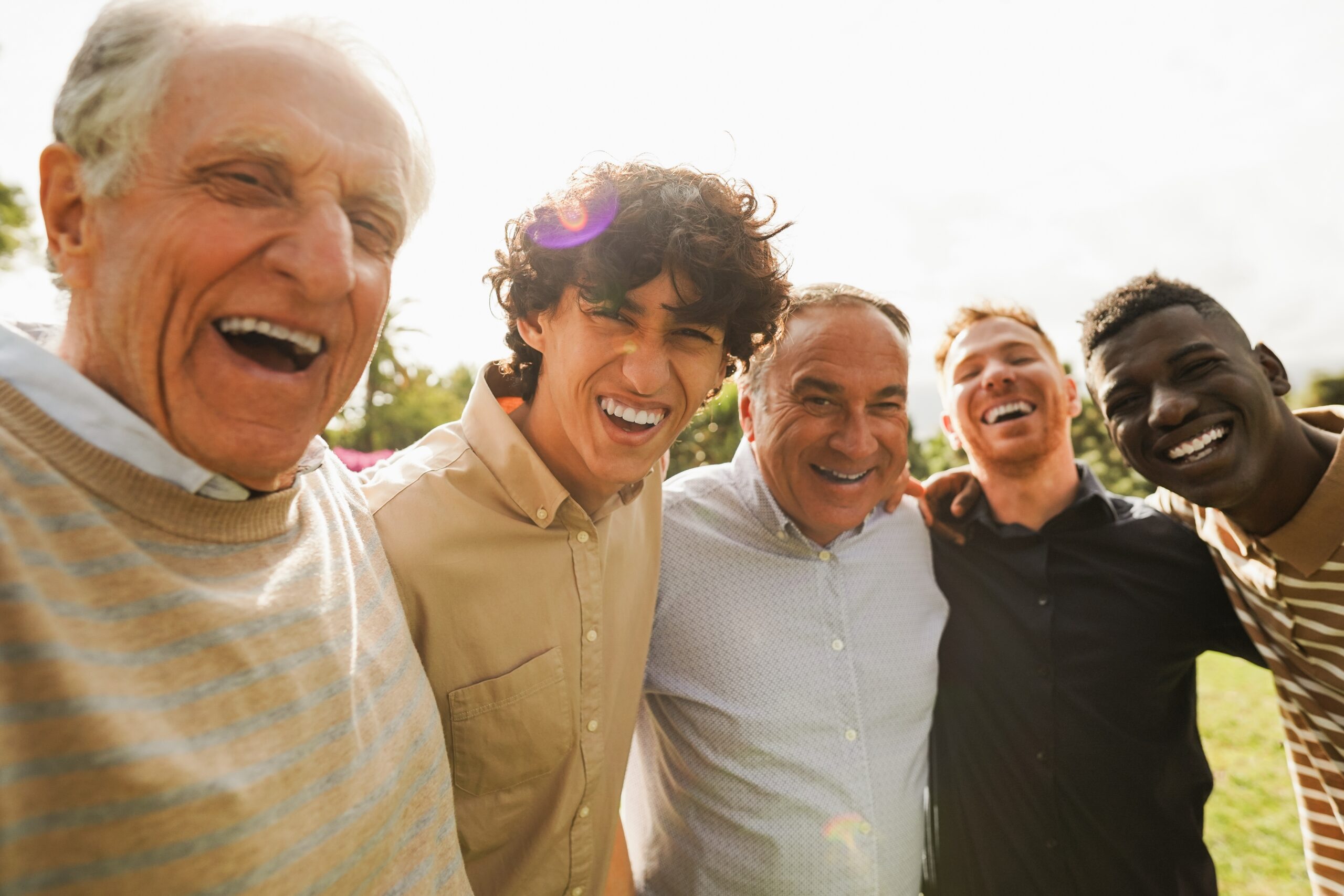 Five diverse men share a joyful moment outdoors, smiling and laughing together in a sunny park setting. Their camaraderie highlights friendship and connection.