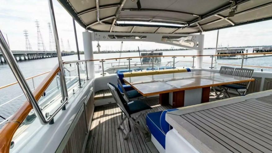 Relaxing deck on a boat featuring a spacious table, seating, and panoramic views of the water and distant power lines, ideal for social gatherings.