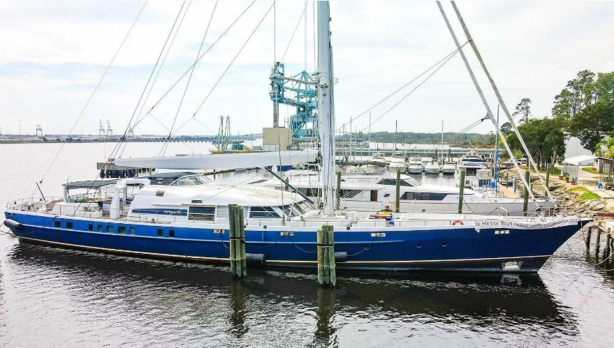 A large blue sailboat docked at a marina with surrounding boats and cranes in the background, showcasing maritime activities.