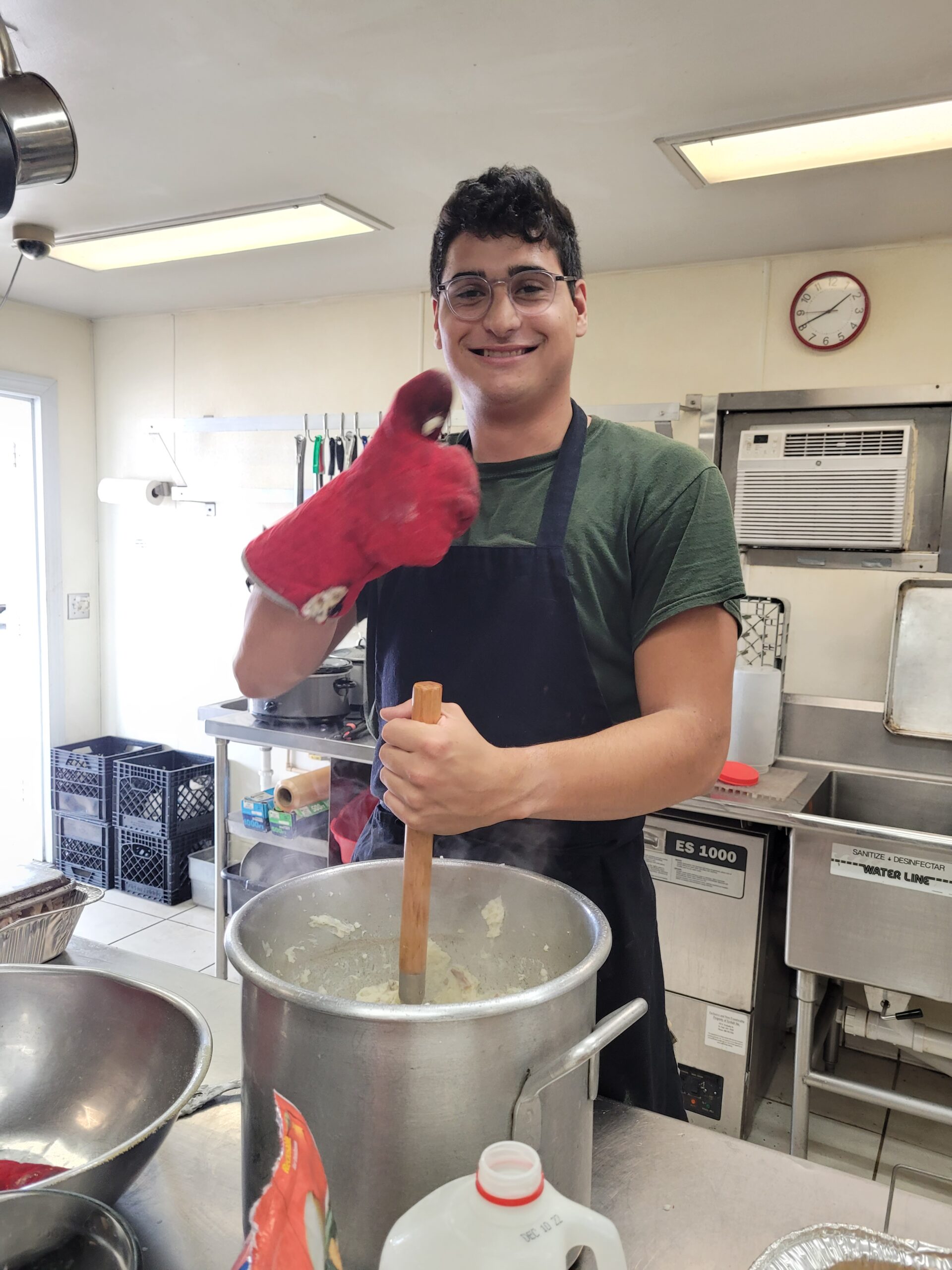 Smiling young man in an apron stirs a large pot of soup in a bright kitchen, wearing an oven mitt and giving a thumbs up.