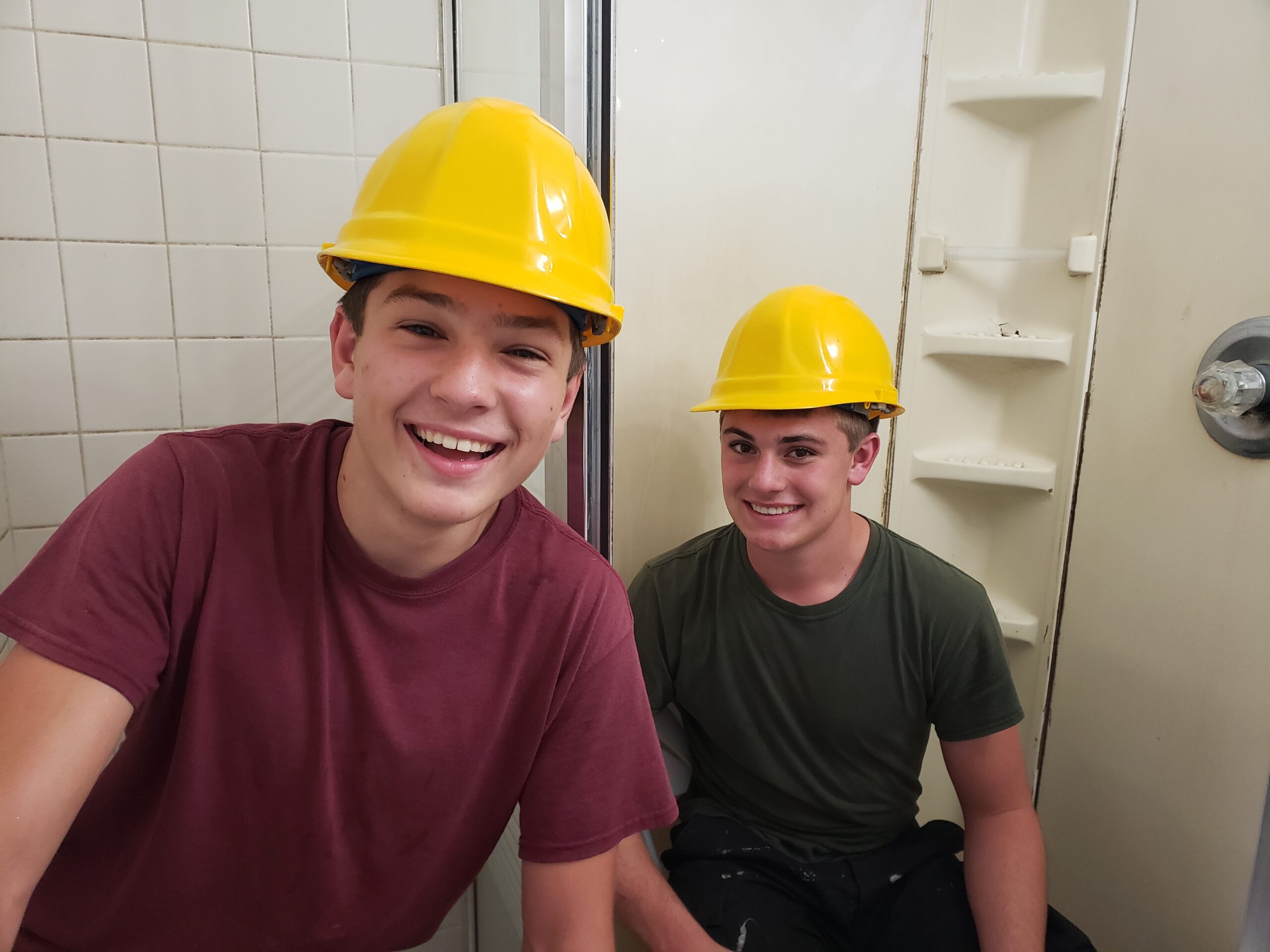Two smiling teenagers wearing yellow hard hats sit in a bathroom, suggesting they are engaged in a renovation or construction project.
