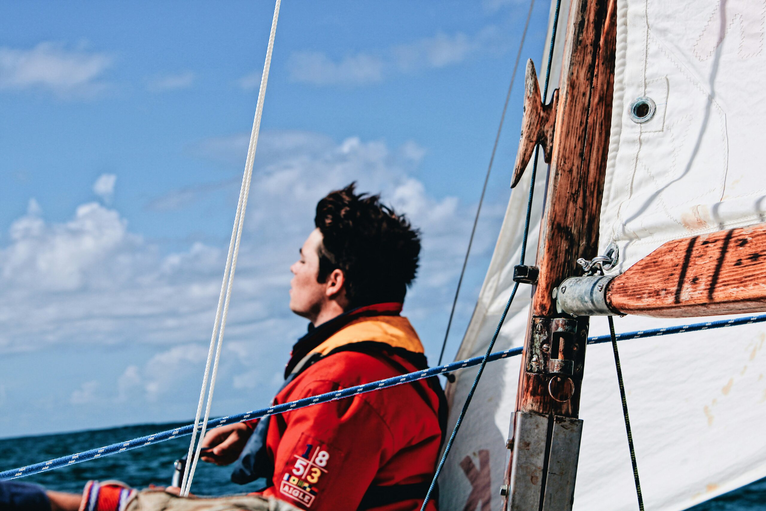 A young sailor in a vibrant orange jacket gazes thoughtfully into the distance as he navigates on a sailboat under a partly cloudy sky.