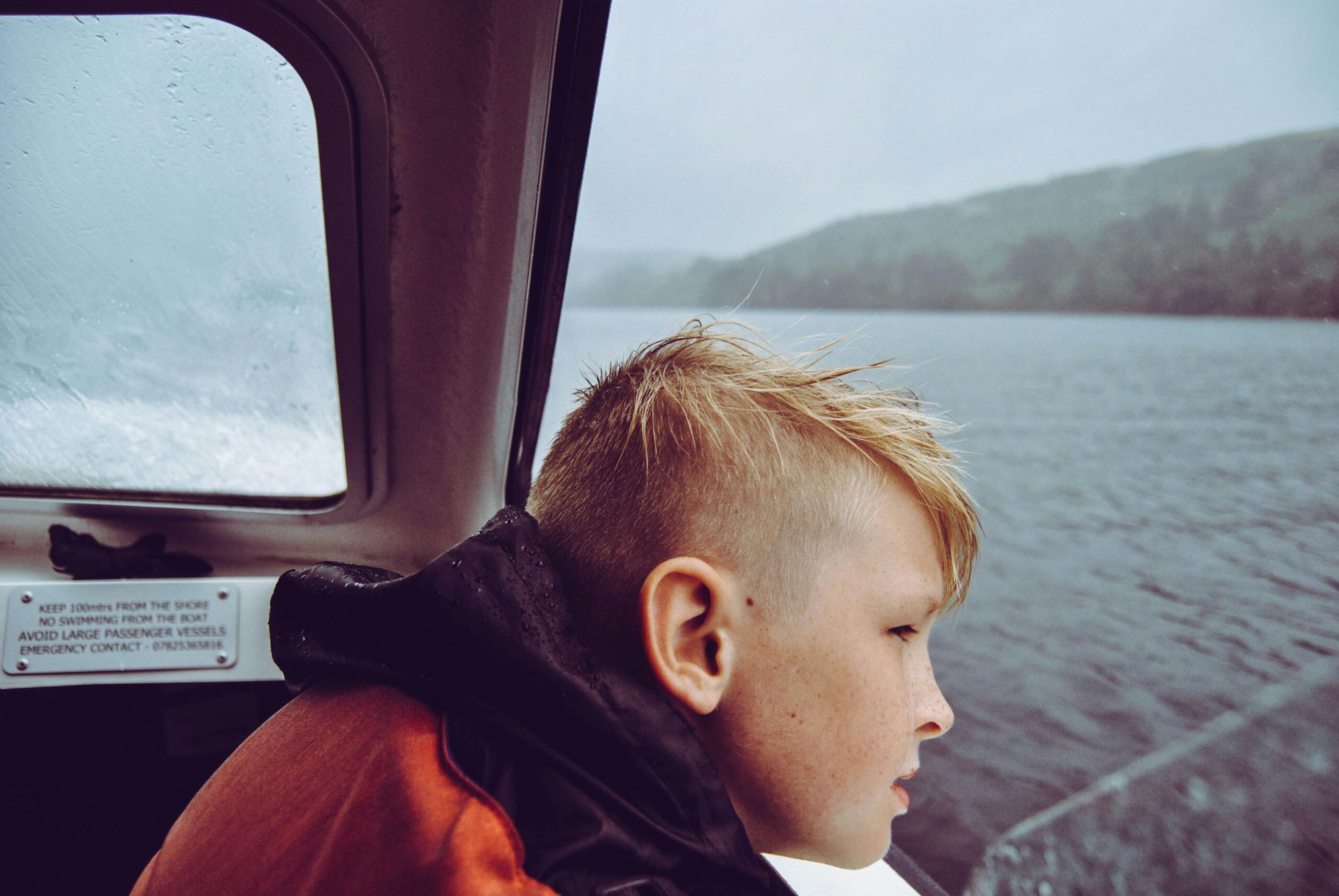 A boy gazes out a boat window onto a rainy lake, reflecting a moment of contemplation amidst a moody landscape.