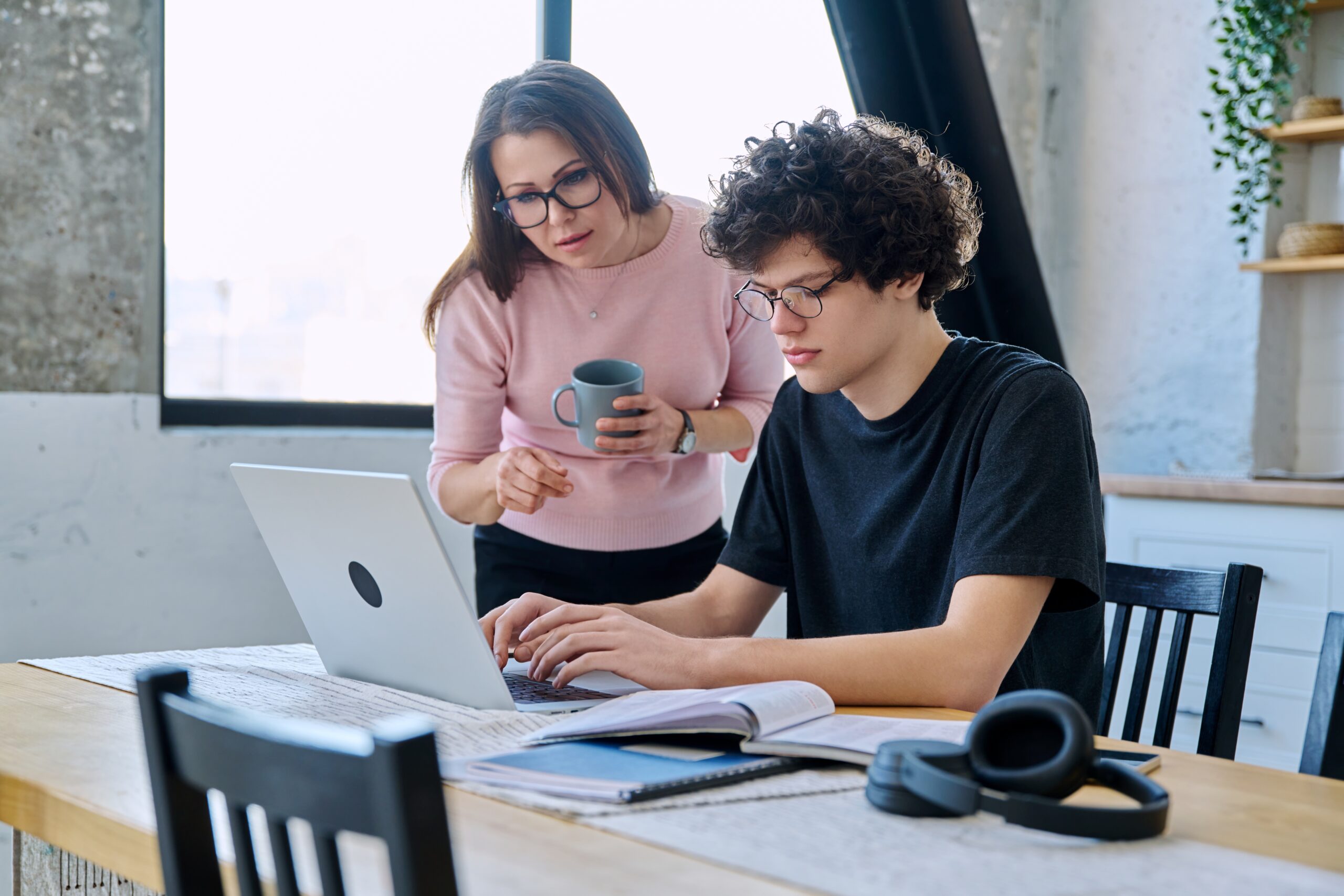 A woman guides a young man using a laptop at a wooden table, surrounded by books and headphones, in a sunlit, modern workspace.