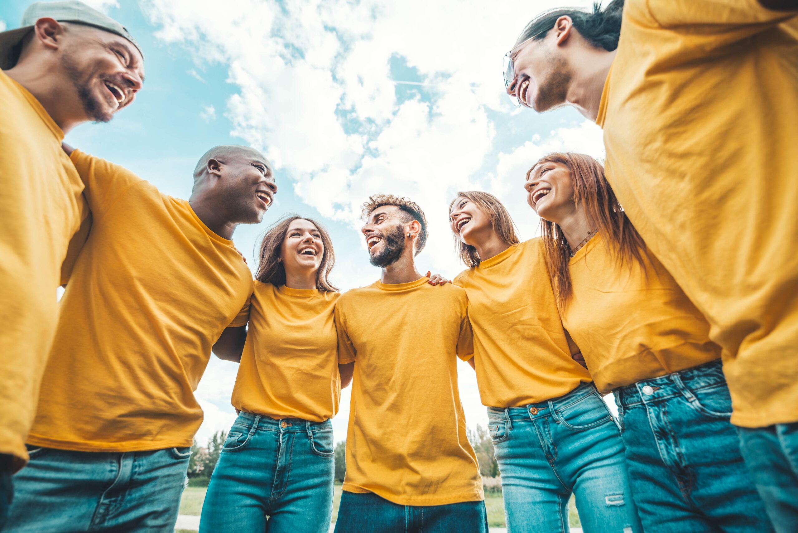 A diverse group of six smiling individuals in matching yellow shirts embrace each other outdoors, radiating joy under a bright blue sky.