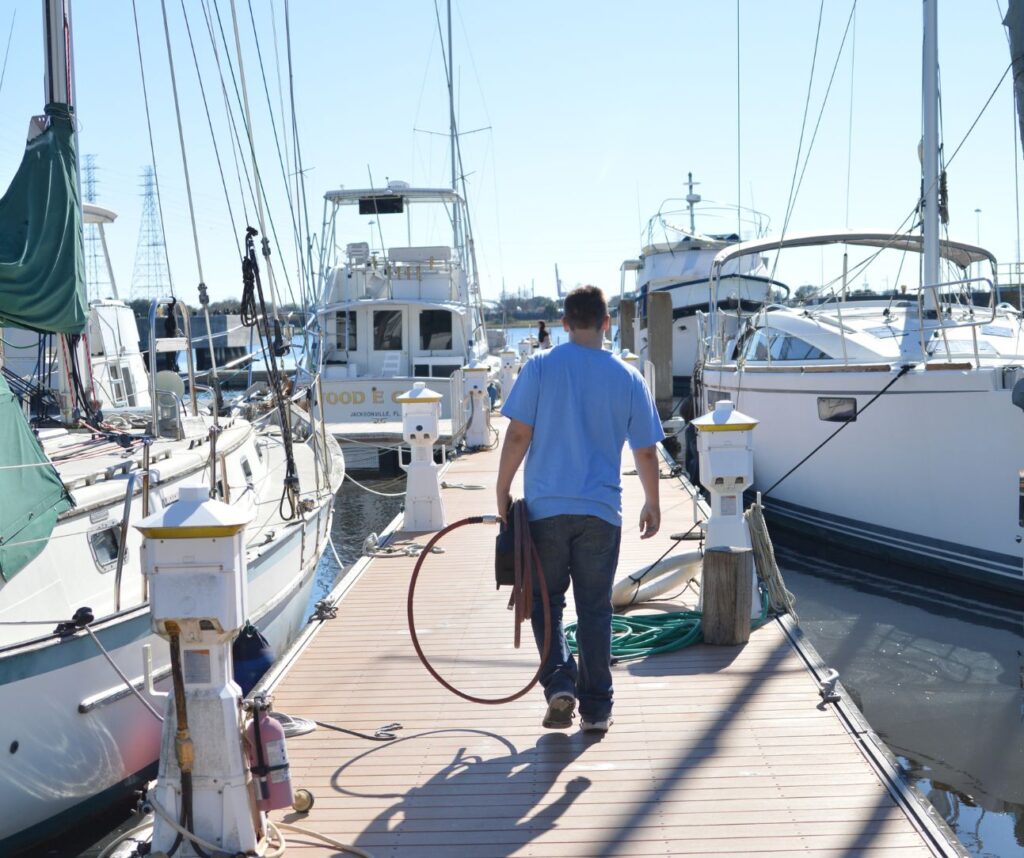 Man in blue shirt walks on a wooden dock between moored boats, holding a red hose and towel. Bright, clear day, marina atmosphere.