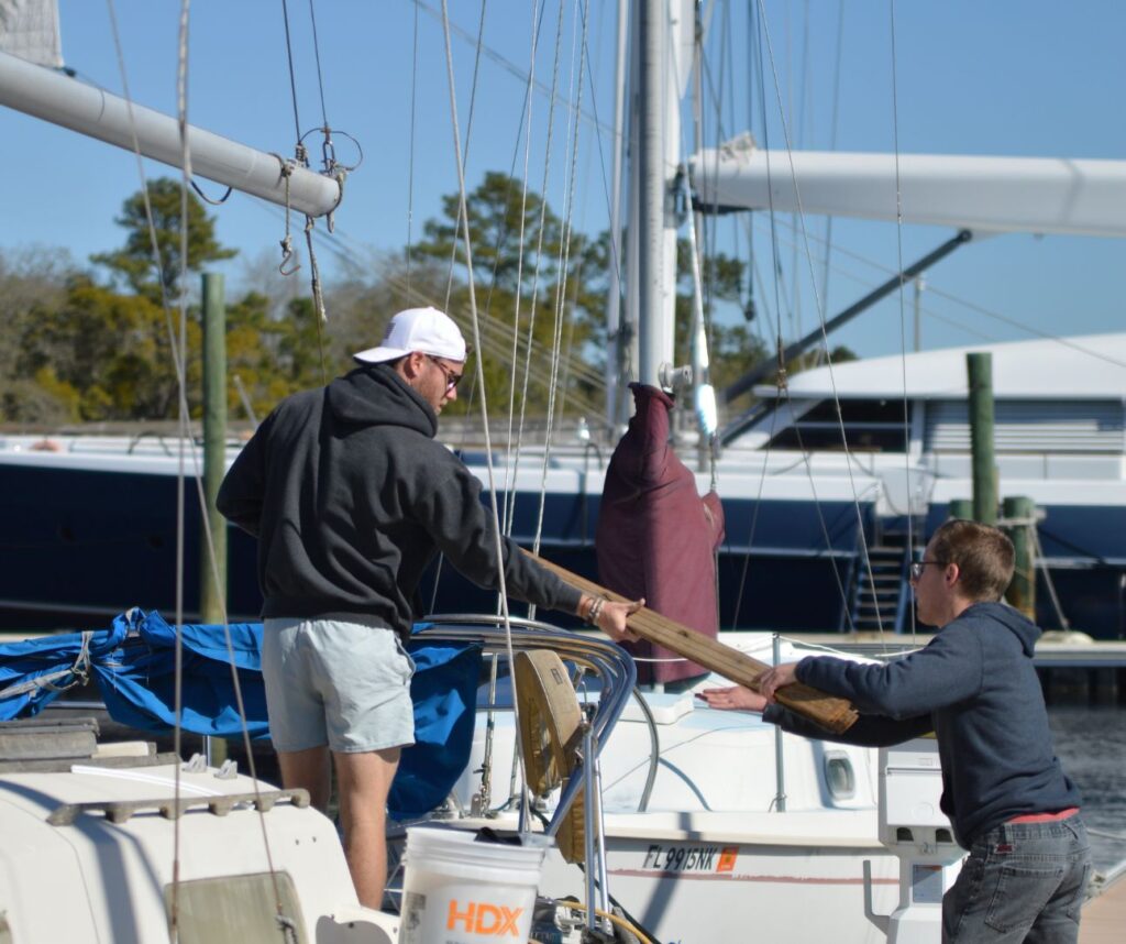 Two men working together on a sailboat docked at a marina. One man hands a wooden plank to the other. The scene is calm and cooperative.